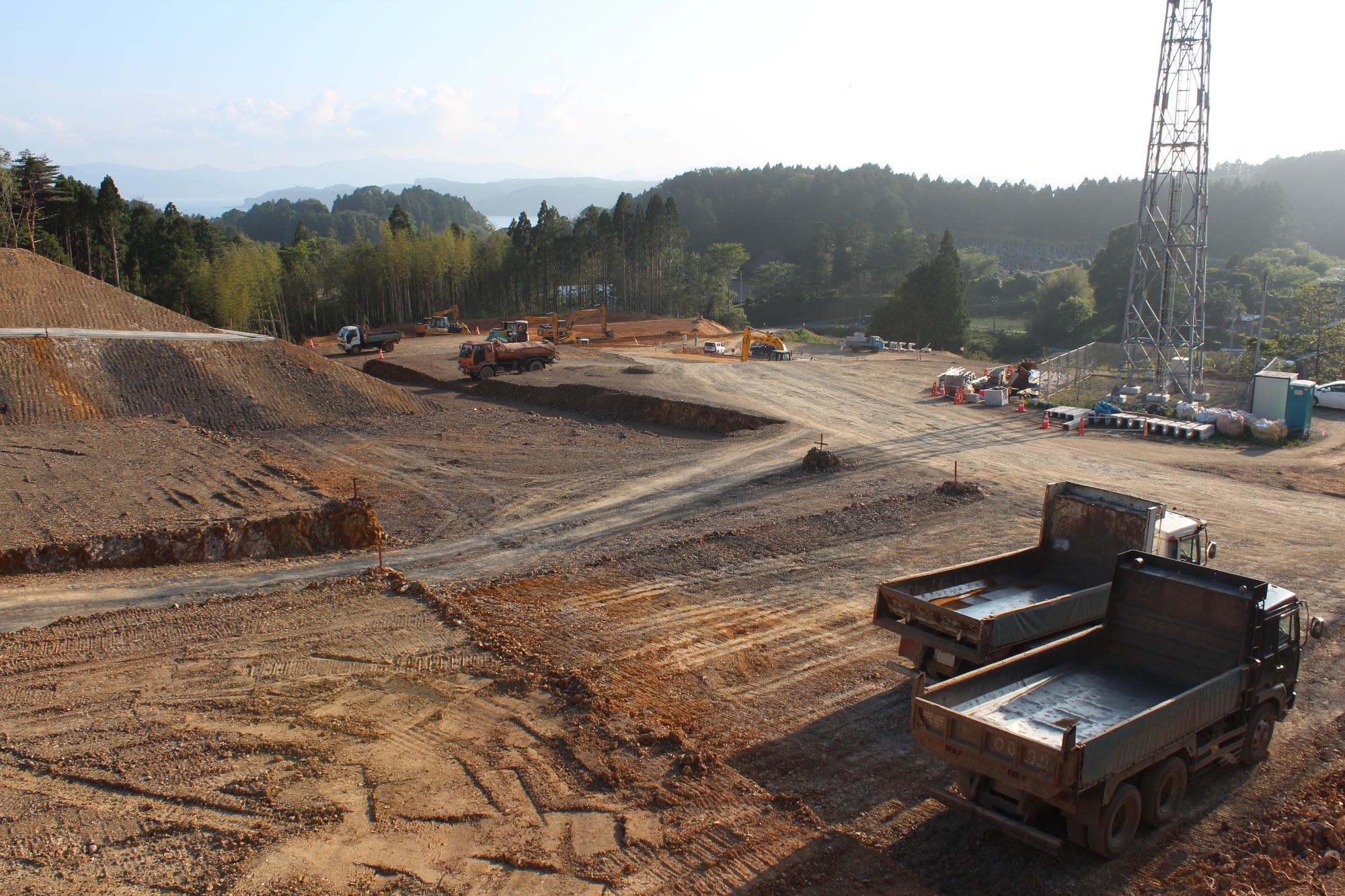 複数のトラックと重機が広い敷地で土地をならしている様子を、高台から見下ろした工事中の様子の写真