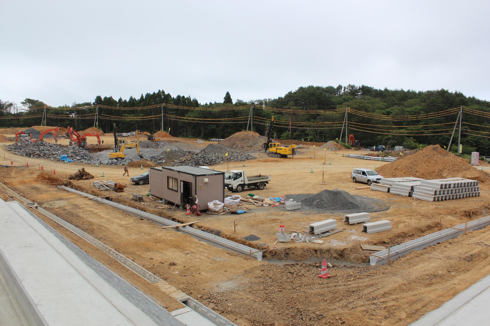 電柱と電線が湾曲しながら空を横切り、複数のショベルカーとトラックが岩や土を運んでいる山あいの造成工事現場の写真
