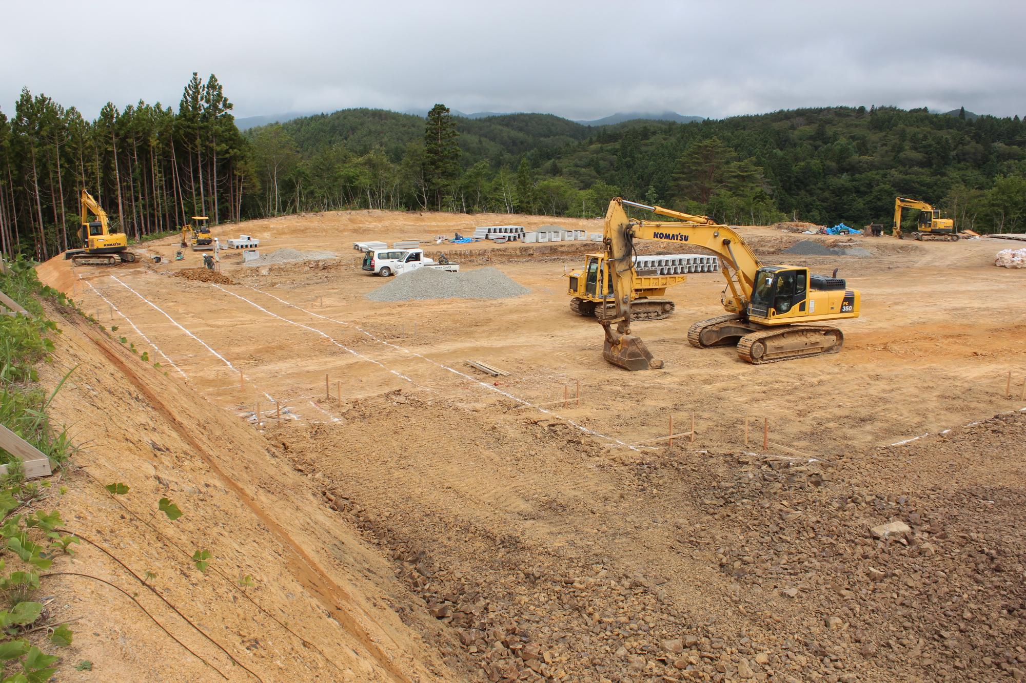 木に囲まれた山あいの平地で、重機やトラックが並び、造成が進んでいる建設予定地の写真