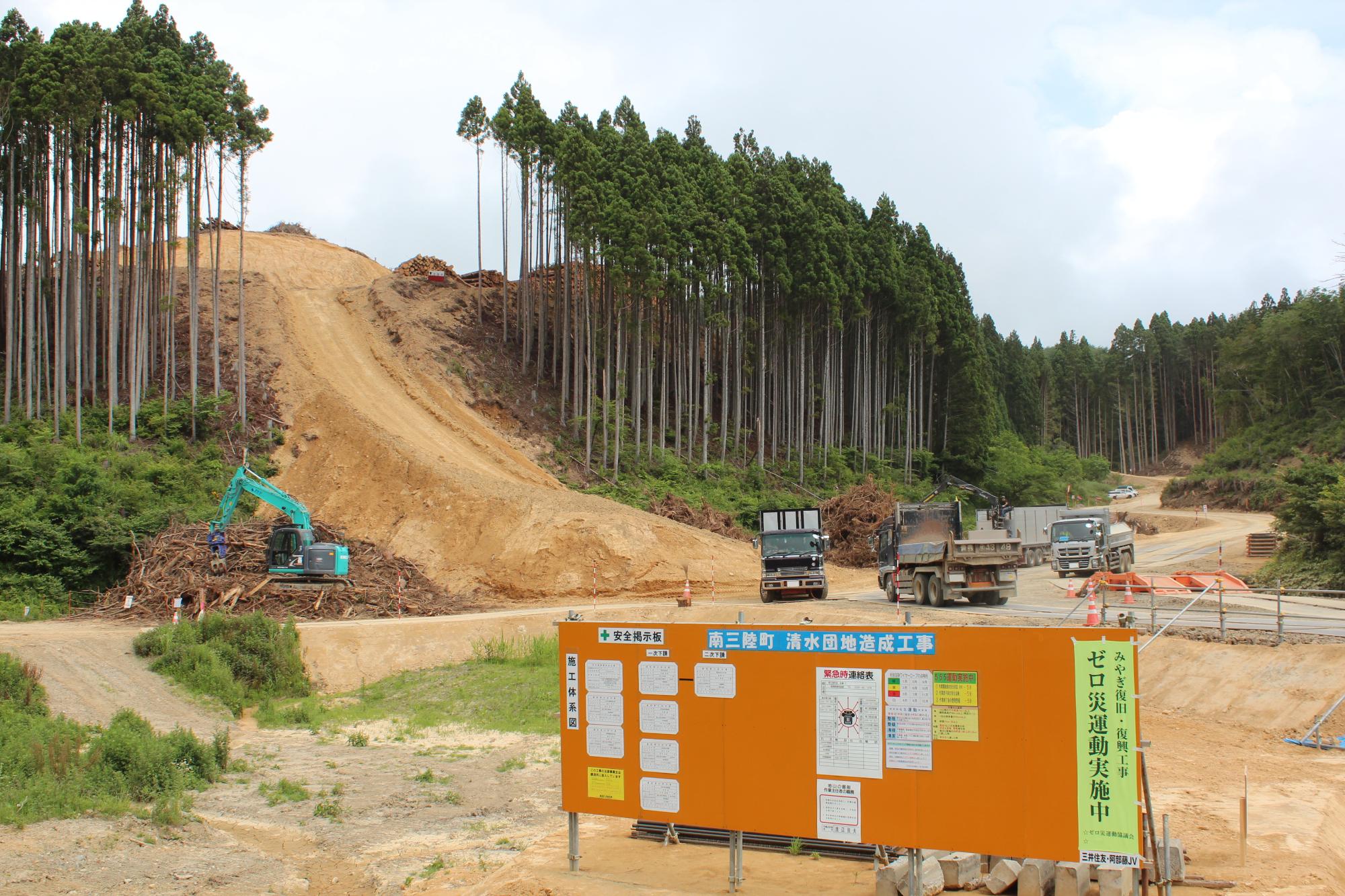 山の斜面を削って道を作るために大型の重機とダンプカーが作業しており、手前に安全掲示板が設置された造成工事の写真