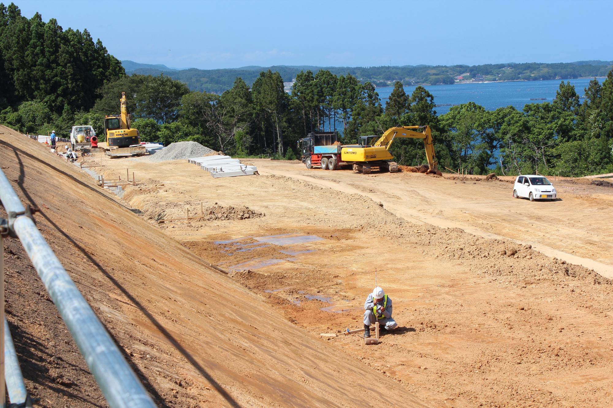 茶色い土の地面が広がった場所に重機や作業車が数台止まり、奥には海が見え、周囲を木々に囲まれている広い工事現場の写真