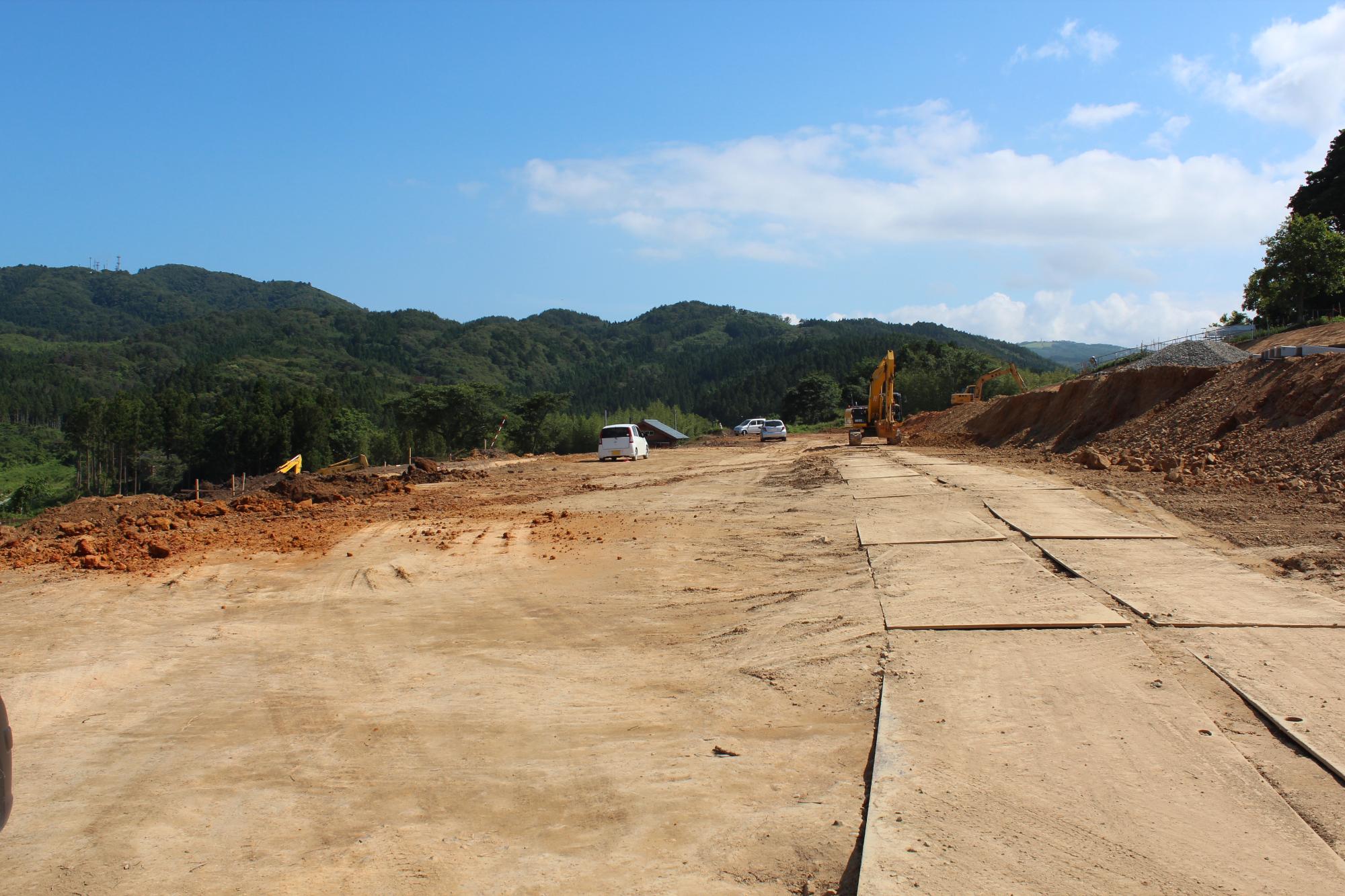 地面に鉄板が敷かれた広い赤土の土地にショベルカーや工事車両が複数見られ、周囲は緑の森林に囲まれている山あいの場所で進められている造成工事現場の写真