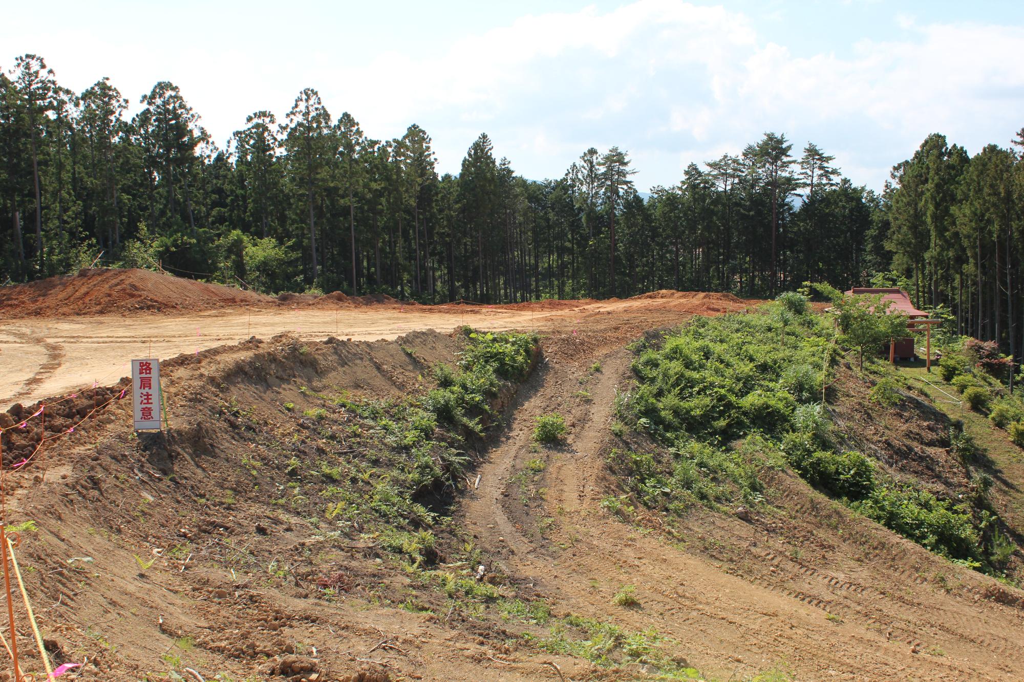 整地が進み、草木が生い茂りはじめた斜面と奥に見える小さな神社、そして「路肩注意」と書かれた看板が設置された高台の写真