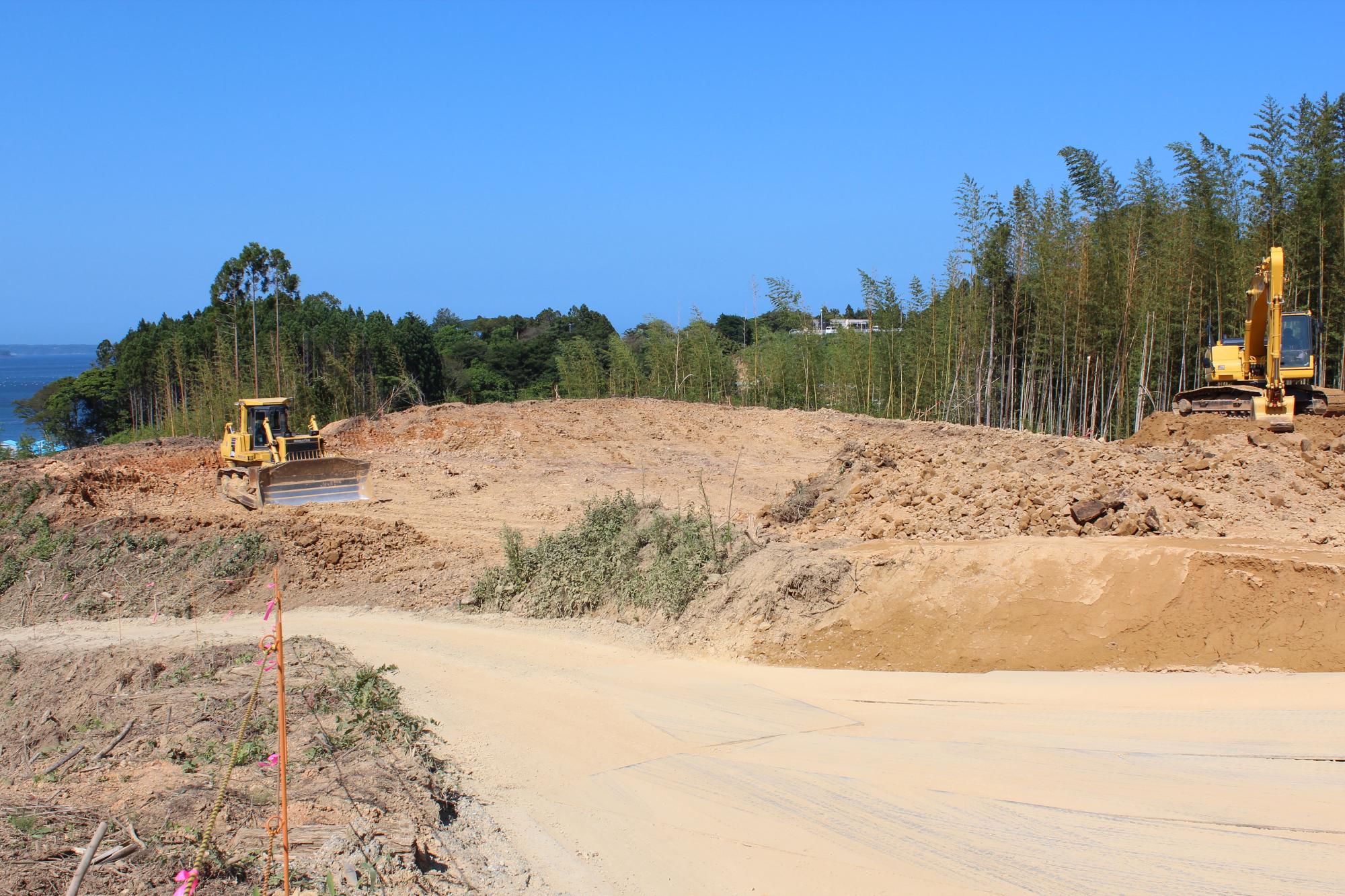 木々に囲まれた高台でブルドーザーとショベルカーが整地作業を進める写真