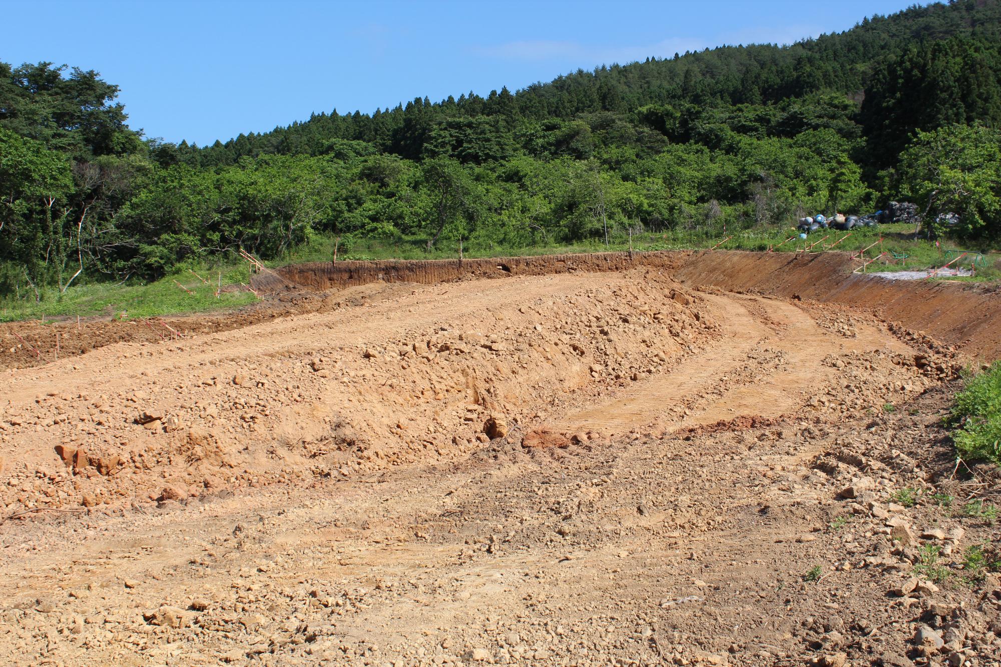 造成工事中の現場で、赤土の道が大きくカーブを描きながら造成された斜面を通り、背景には濃い緑に覆われた山林が広がっている写真