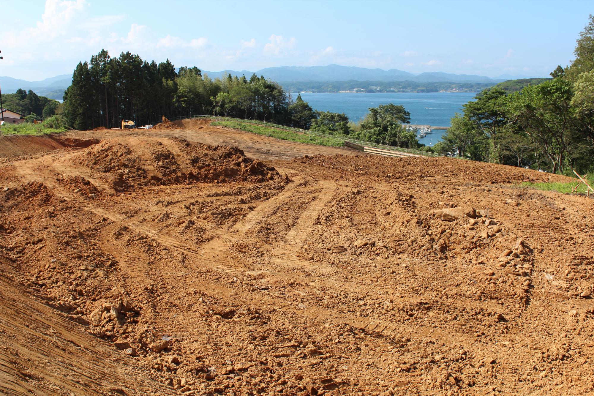 造成工事が進み、赤土がむき出しになった広い斜面に重機の跡がくっきりと残り、その奥には濃い緑の林と青く広がる海、その向こうに山々が連なっている写真