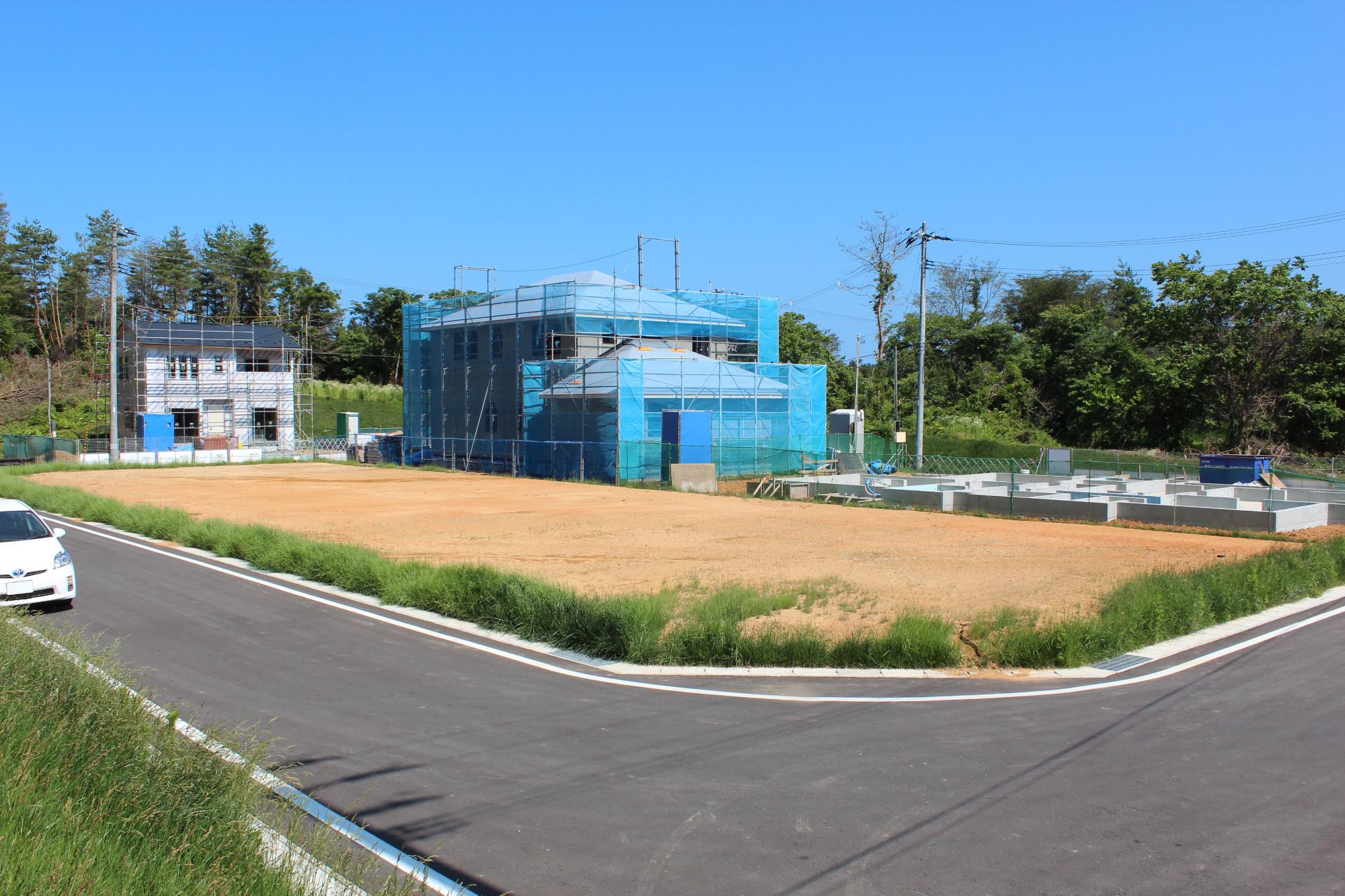 青空の下、緩やかにカーブする新しい舗装道路に沿って草が生い茂り、その奥には整地された土の区画が広がり、ブルーの防護ネットに覆われた複数の建設中の住宅やコンクリート基礎が整然と並んでいる様子を捉えた写真