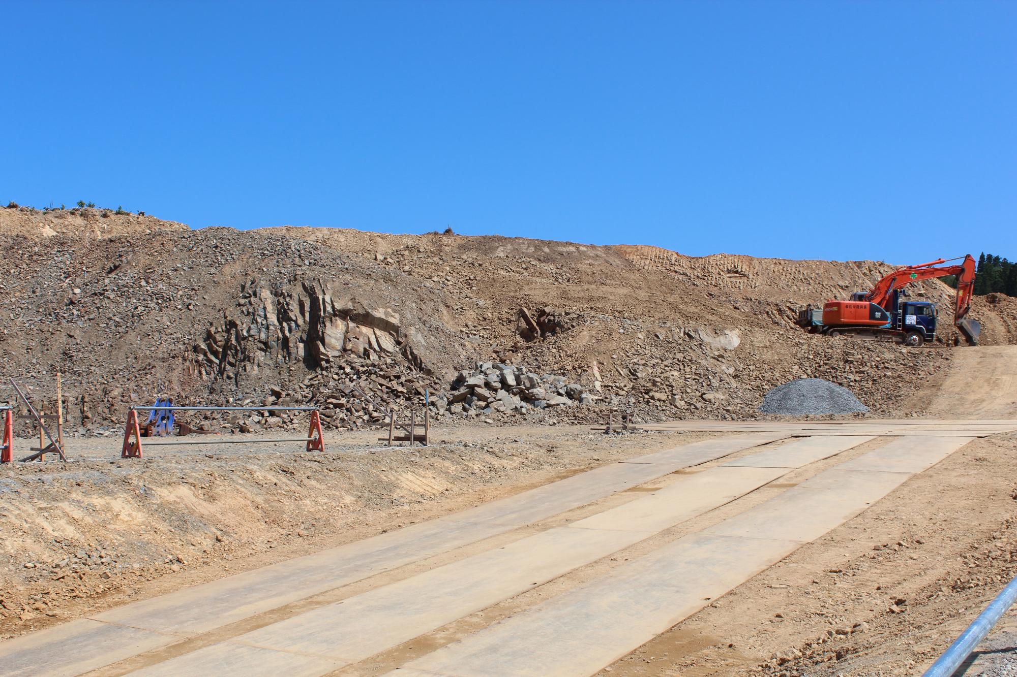 青空の下、重機が岩を削って整地作業を行っている広大な造成工事現場で、削られた岩や土が山のように積まれ、中央には仮設の鉄板道路が敷かれている写真