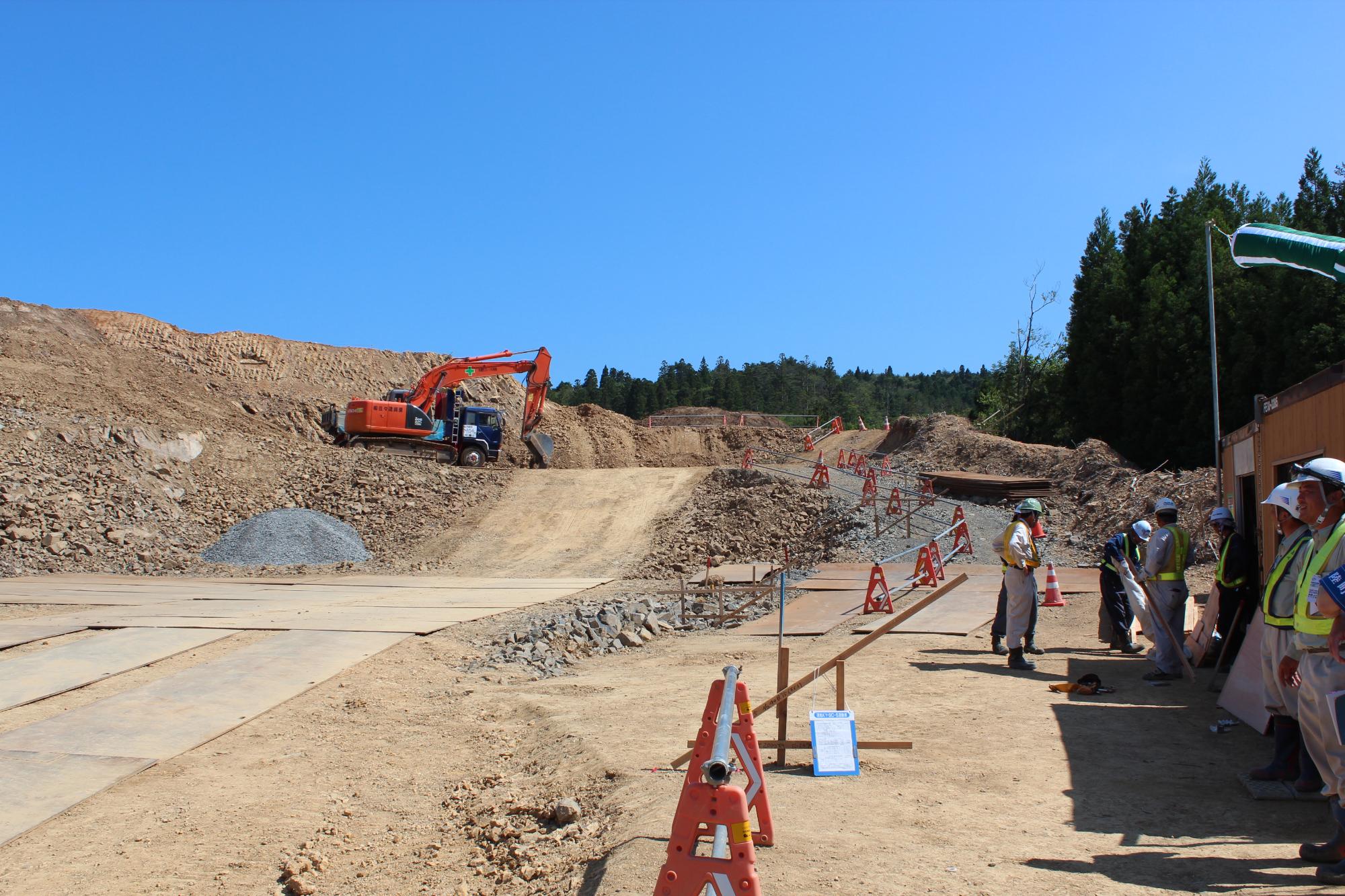 青空の下、重機が稼働する造成工事現場で、土砂が削られた斜面に沿って仮設の通路が整備され、多数の作業員たちが安全ベストとヘルメットを着用して作業打合せをしている様子の写真