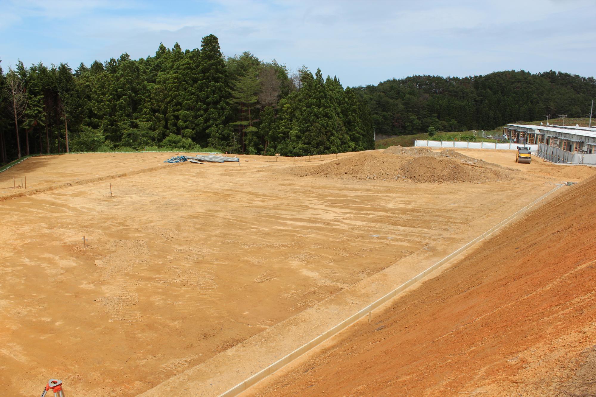 造成が終わった広い敷地に土の山があり、奥に建物と機械が見える写真