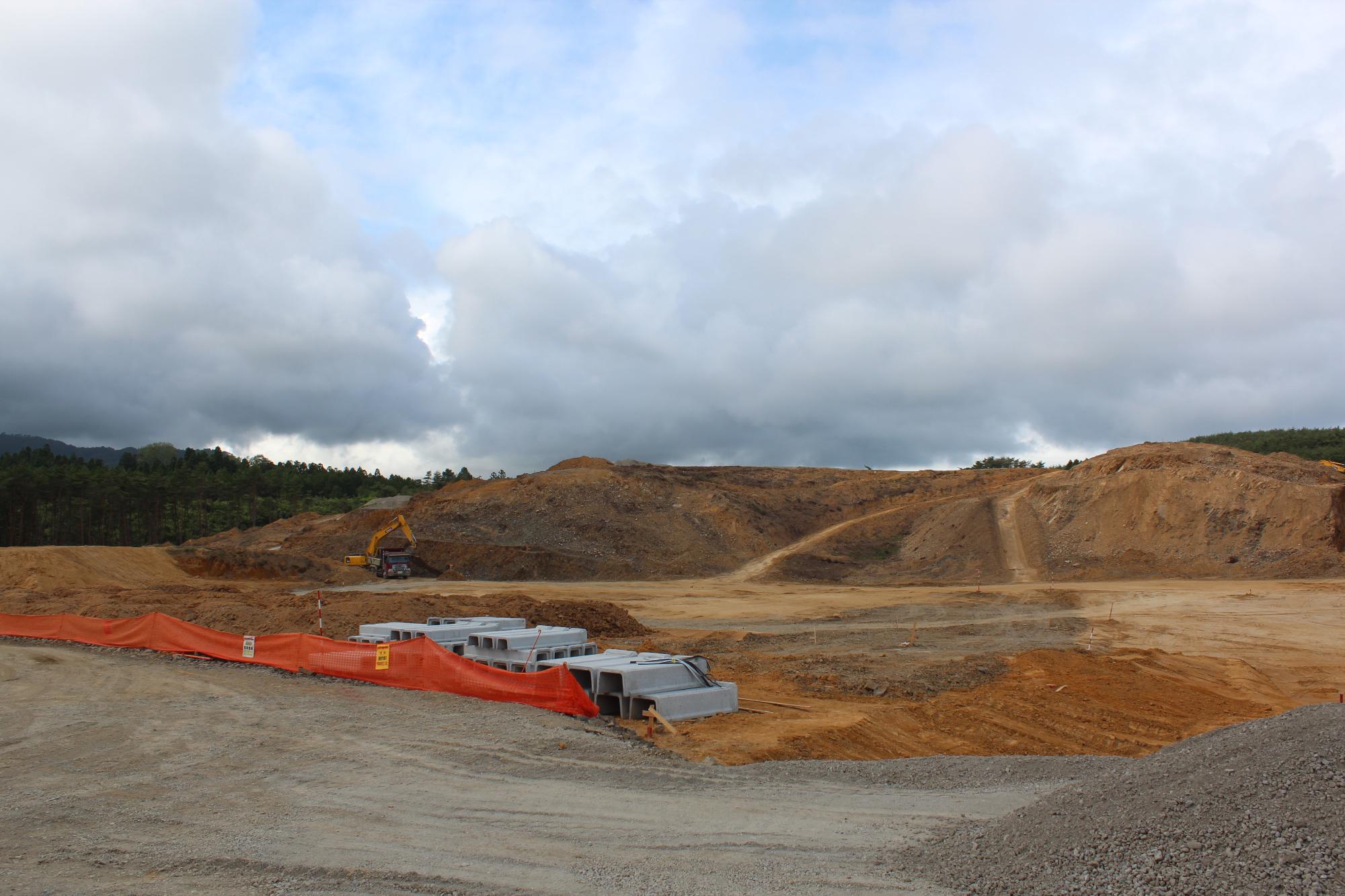 広大な造成地で重機が稼働しており、オレンジ色のフェンスとコンクリート資材が整然と並ぶ、曇り空の下での工事現場の様子の写真