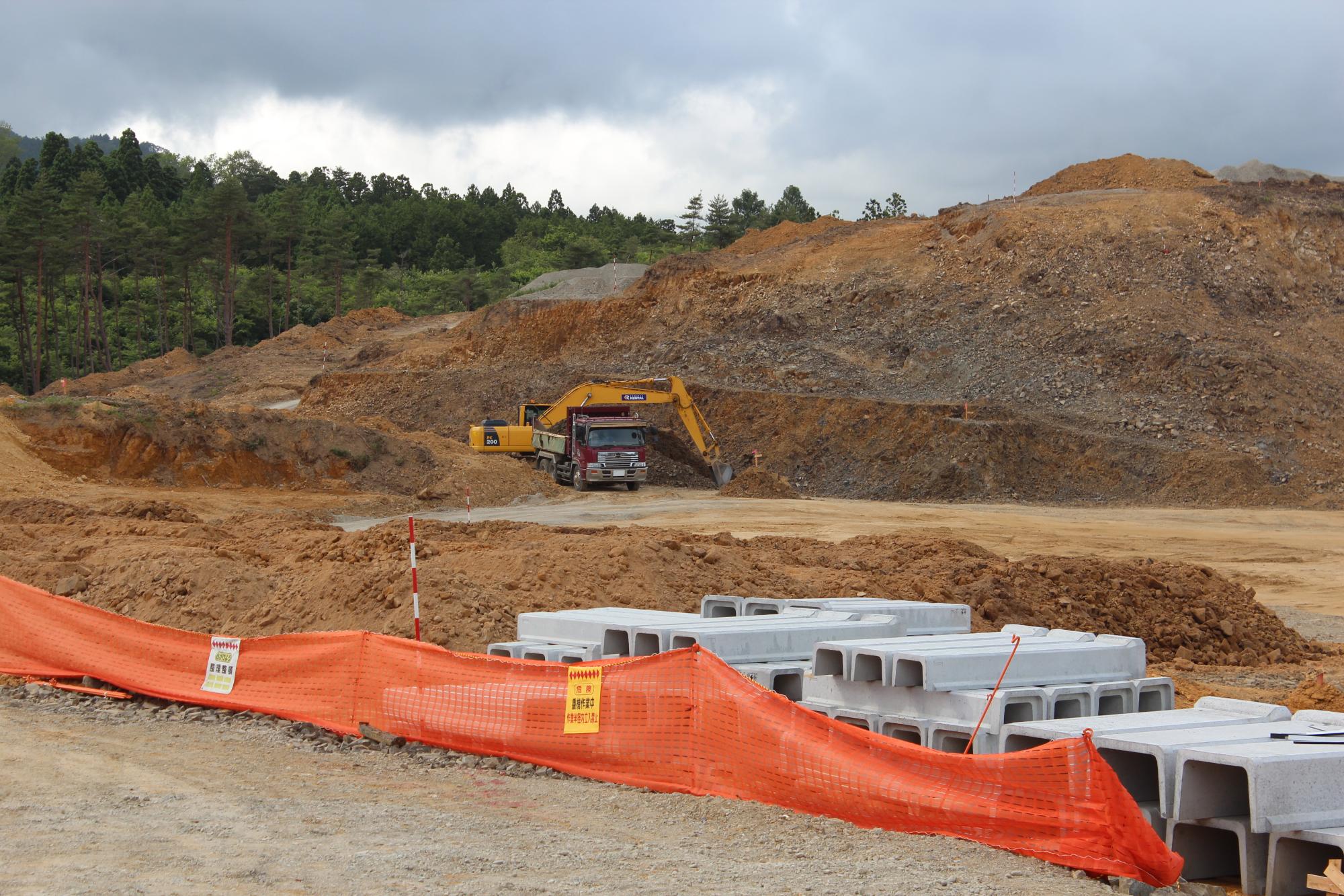 森林の前で、ショベルカーが大型ダンプに土砂を積んでいる造成地の風景で、手前にコンクリート資材とオレンジ色の安全フェンスがある工事現場の写真