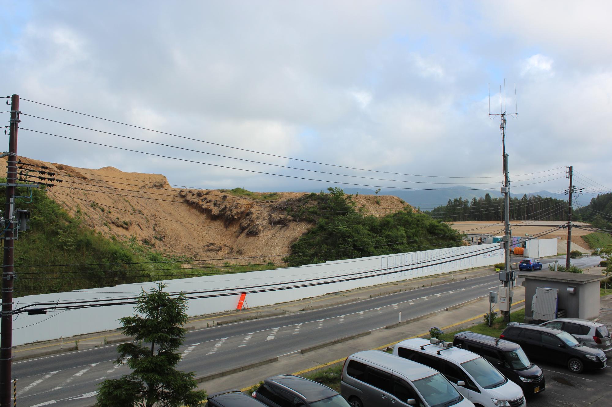舗装された道路の向こう側に白い工事用フェンスと造成中の土の斜面が広がり、その手前には駐車中の車と電柱が立ち並ぶ様子を捉えた写真