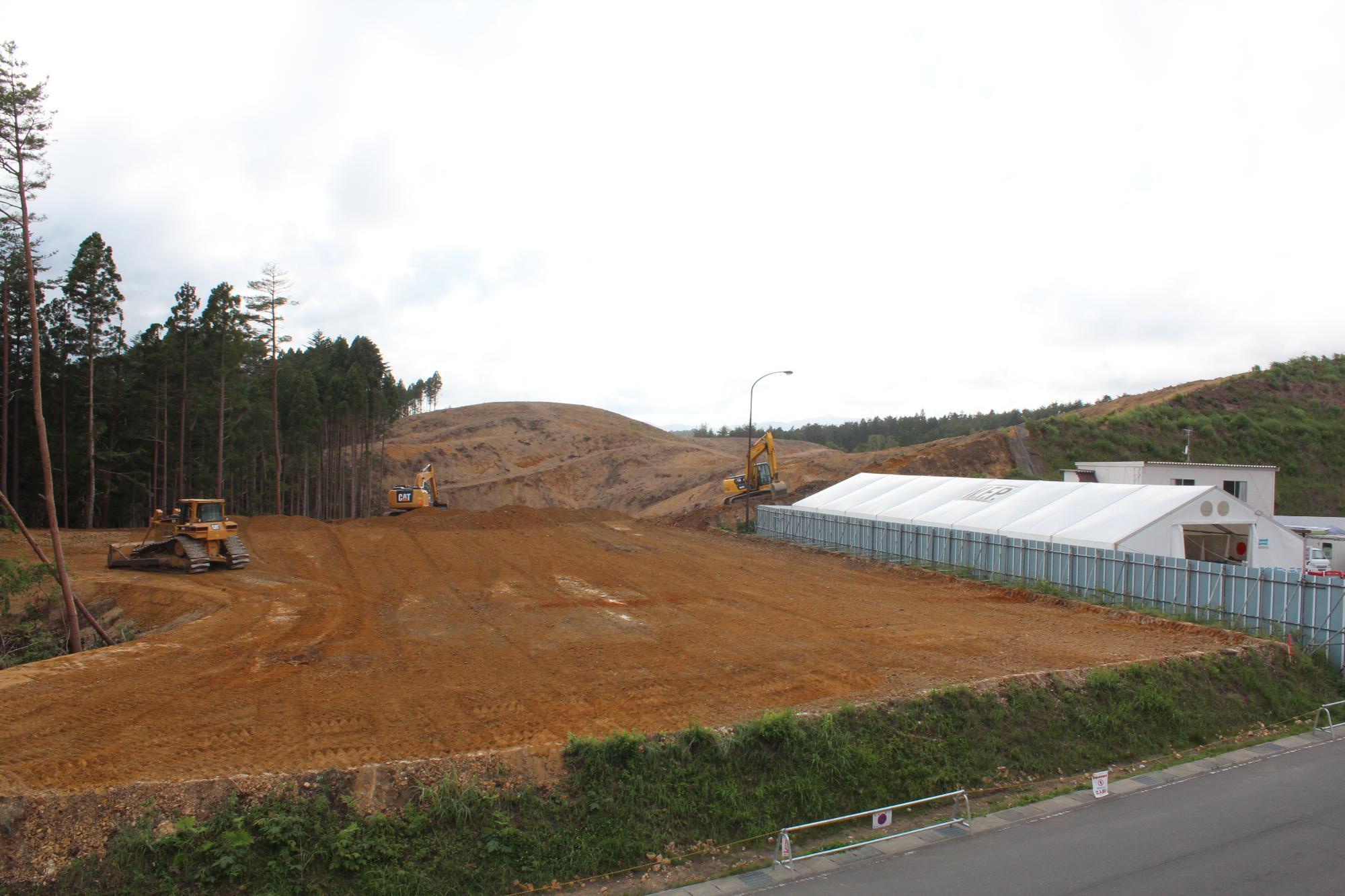 造成工事中の広い赤土の土地にブルドーザーやショベルカーが並び、隣には仮設の白いテント建物と緑の斜面が見える写真