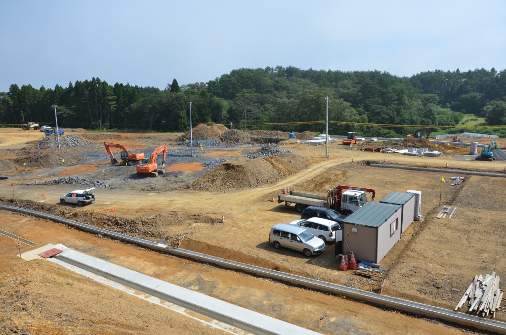 林に囲まれた広大な敷地で、仮設事務所や小型トラック、コンクリート製の資材が点在し、複数の重機が岩を掘り起こしている造成工事の途中の写真