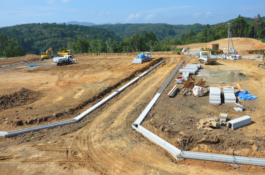 複数の重機とトラックが入り、U字溝や資材が並ぶ造成工事が進行中の広い土地の写真