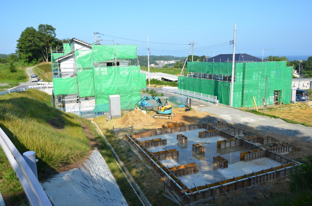 造成地の一角で建設途中の家々が緑のシートで覆われ、手前には住宅の基礎工事が進んでいる写真