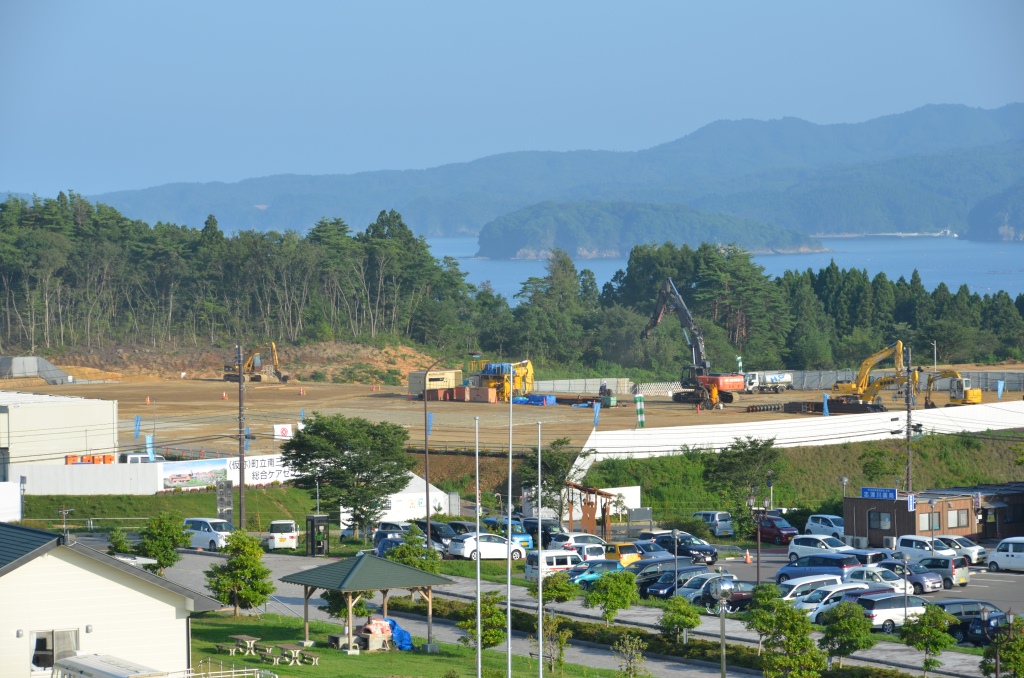 周りを白いフェンスのような物で囲まれた造成地に複数のショベルカーが配置され、その奥に海と山が広がっている写真