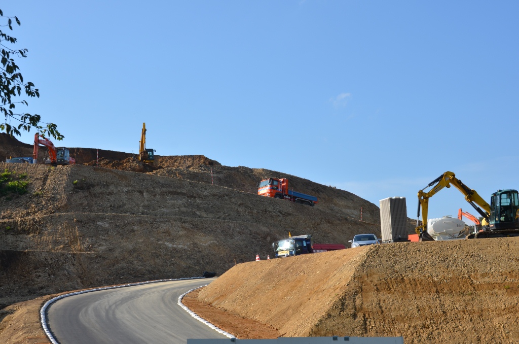 急勾配の山を切り崩しながら重機やトラックが工事を進めている、舗装された道路が見える写真