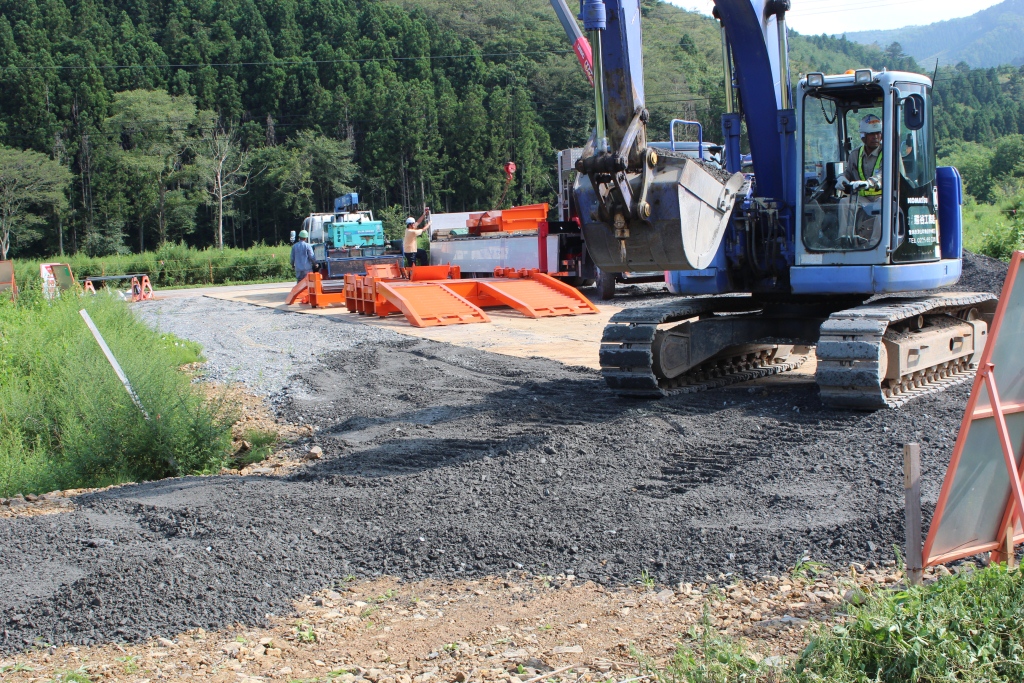 手前にショベルカーを操縦している作業員、奥に重機のそばで作業している作業員やオレンジ色の仮設スロープや資材が見える周囲を木々や草に囲まれた作業現場の写真