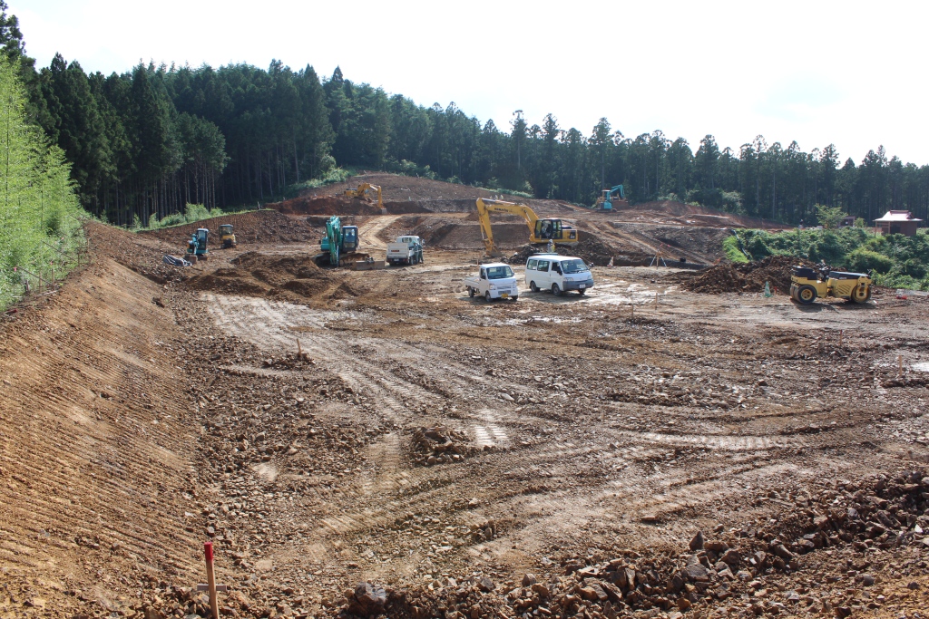 森林を切り開いて整地された広い工事現場で、複数の重機や作業車が作業をしている様子の写真