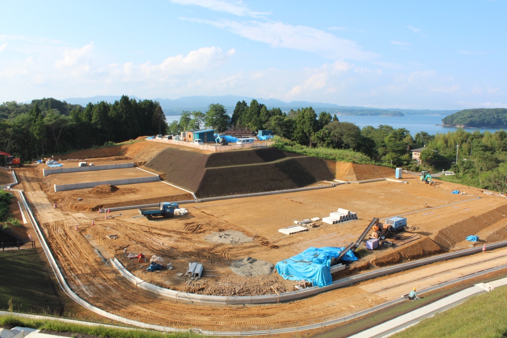 段々に整地された造成地で工事車両が作業を進め、奥には海と緑が広がる風景が見える写真