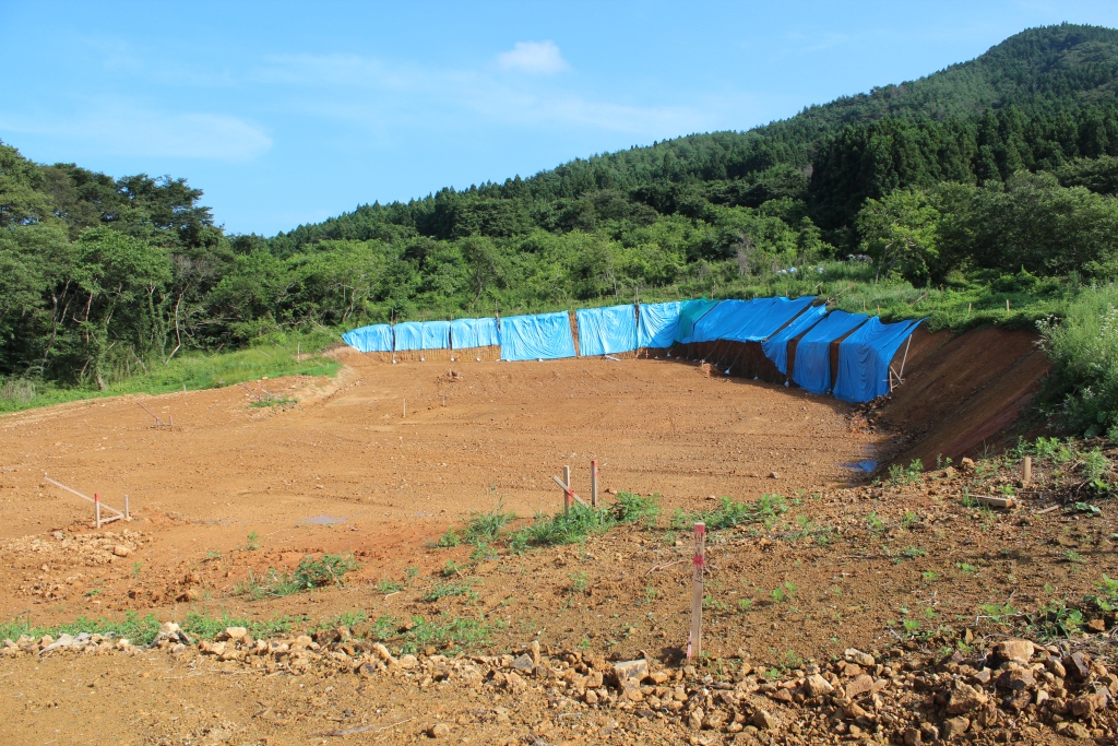 森林に囲まれた山間の造成地で、地面は赤土でならされ、右奥の斜面には土砂崩れを防ぐための青いシートが丁寧にかけられ、ところどころに工事用の杭が立っている写真