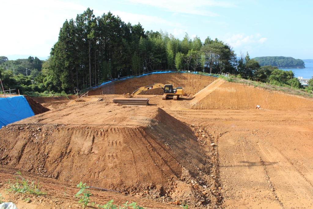 造成工事が進む赤土の広い土地で、中央に作業中のショベルカー、背後には竹や杉の木が並ぶ林と海が見える写真