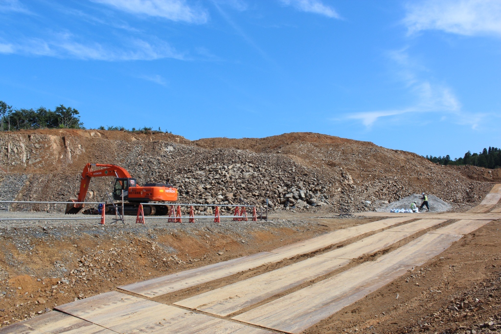 山を切り崩して造成が進む現場で、中央にはオレンジ色のショベルカーが停止しており、右奥では作業員が砂利の山の前で作業をしている様子が見える写真