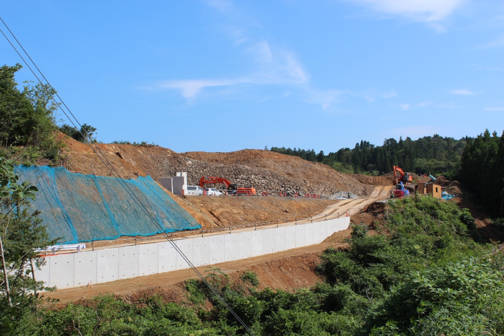 山を切り開いて造成が進められている現場で、左側の斜面にはブルーシートがかけられ、中央にはコンクリートの擁壁が築かれ、奥では重機が稼働している様子を捉えた写真