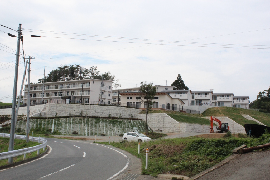 丘の上に建つ白い集合住宅群とその手前に工事用重機と白い車が停まっている様子をカーブした道路越しに見上げた風景の写真
