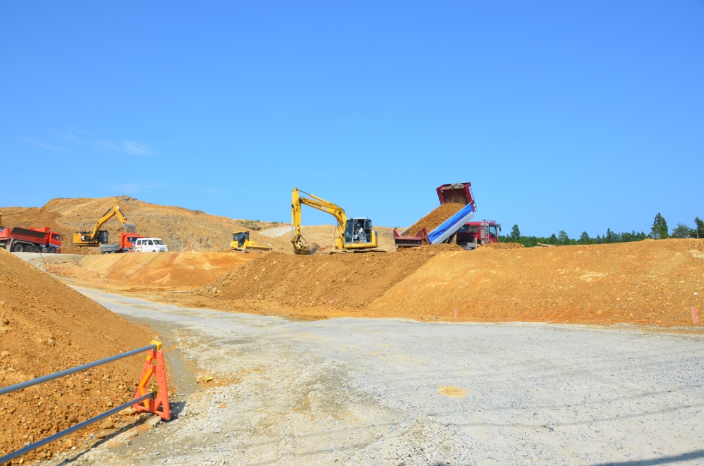 鮮やかな青空の下、山の中腹で重機やダンプカーが土砂を運び出している造成工事の様子の写真