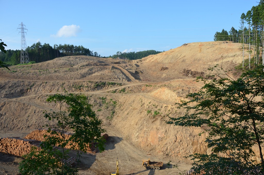 山の斜面が大規模に切り開かれた造成地で、背景に残された森林と送電線の鉄塔が立っている写真