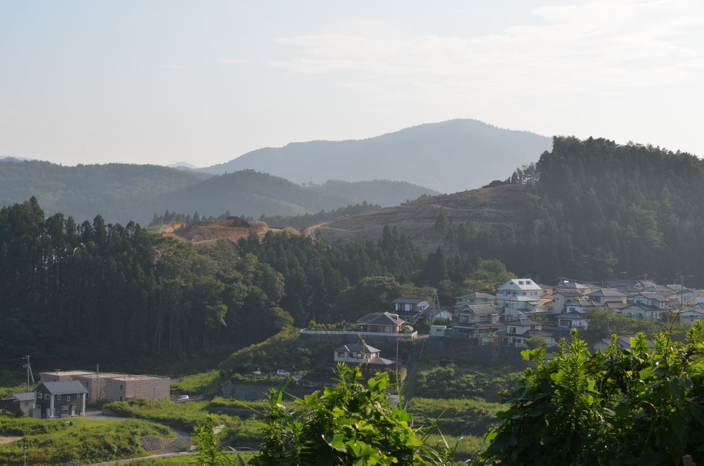伐採が進んだ山の中腹に広がる赤土の造成地と、それを見下ろすように手前に立ち並ぶ住宅街が広がり、背景には緑深い山並みが連なっている写真
