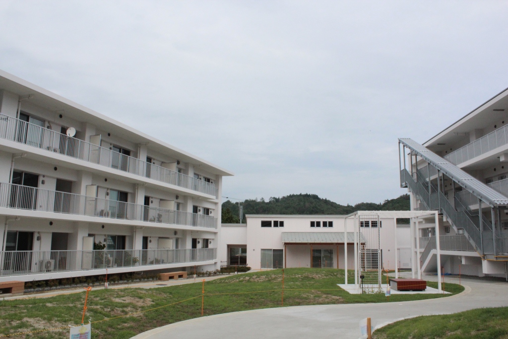 白い外壁の3階建て集合住宅と低層の建物が芝生の広場を囲むように配置された住宅団地の写真