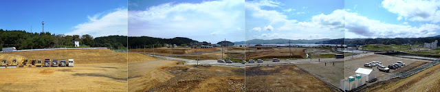 高台から広がる海や山が見渡せる復興工事エリアを見下ろし、整地された土地や車両が並ぶ様子をとらえたパノラマ写真