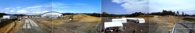 左奥にドーム型の屋根の体育館のような建物が建つ広大な敷地で工事が進行中で、手前にはテントや見学者が写るパノラマ写真