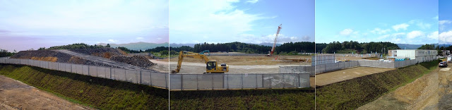フェンスで囲まれた広大な敷地内で重機が作業しており、土が盛られた山や広場が広がるパノラマ写真