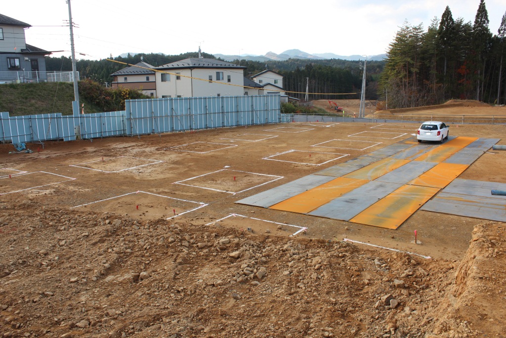 オレンジ色の鉄板が敷かれた造成地に車両が入り、奥では工事車両や作業員が活動している林に囲まれた現場の写真