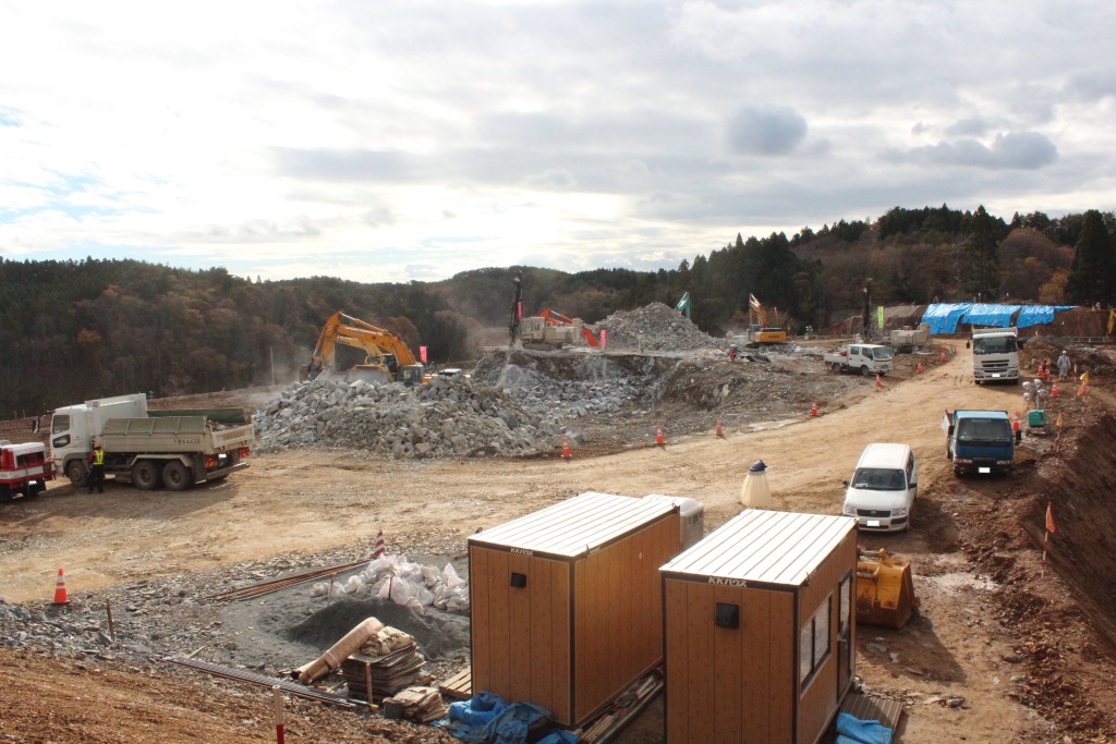 砕石が山のように積まれた造成現場で、ダンプカーや重機が作業中の様子を上から撮影した工事の写真