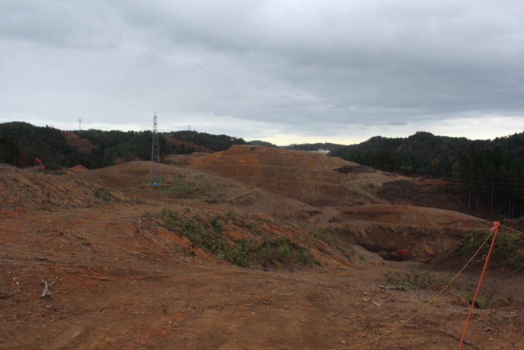 山あいの広大な土地が重機によって大きく造成され、地面が削られて起伏が激しい赤土の風景が広がり、ところどころに重機や送電線の鉄塔が点在し、遠くに山並みと曇り空が見える写真