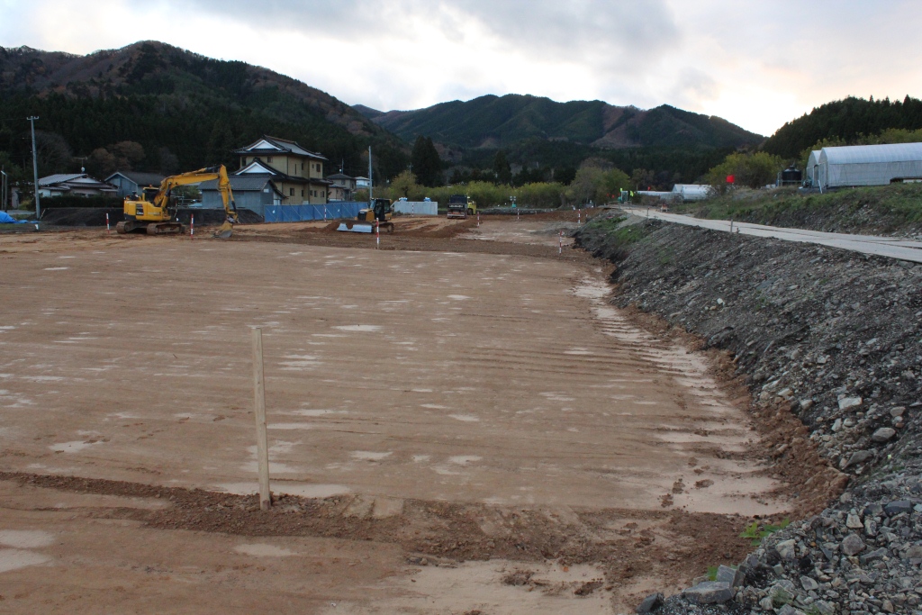 広く整地された土地に作業中のショベルカーがあり、土地の奥には瓦屋根の日本家屋、右手にはビニールハウスや細い舗装道路、背景には山々が連なっている写真