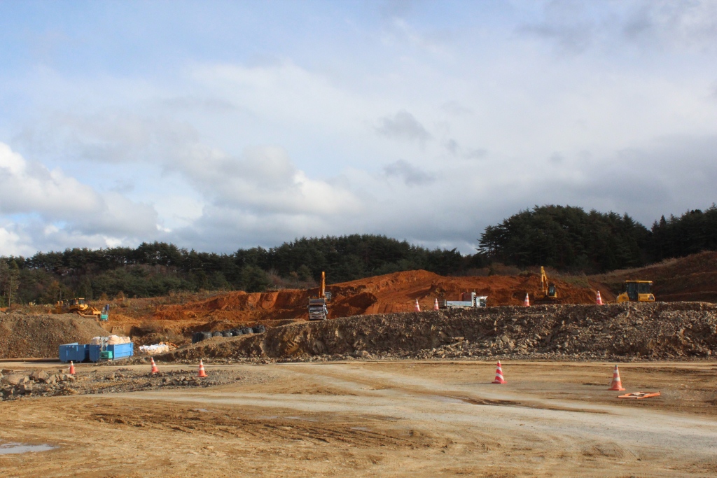 重機とダンプカーが赤土の斜面を整地している山間部の工事現場の写真