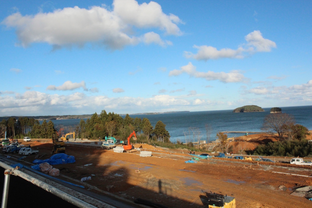 小さな島と青い空が広がっている海のそばで重機やトラックが多数使われている大規模な工事現場の写真