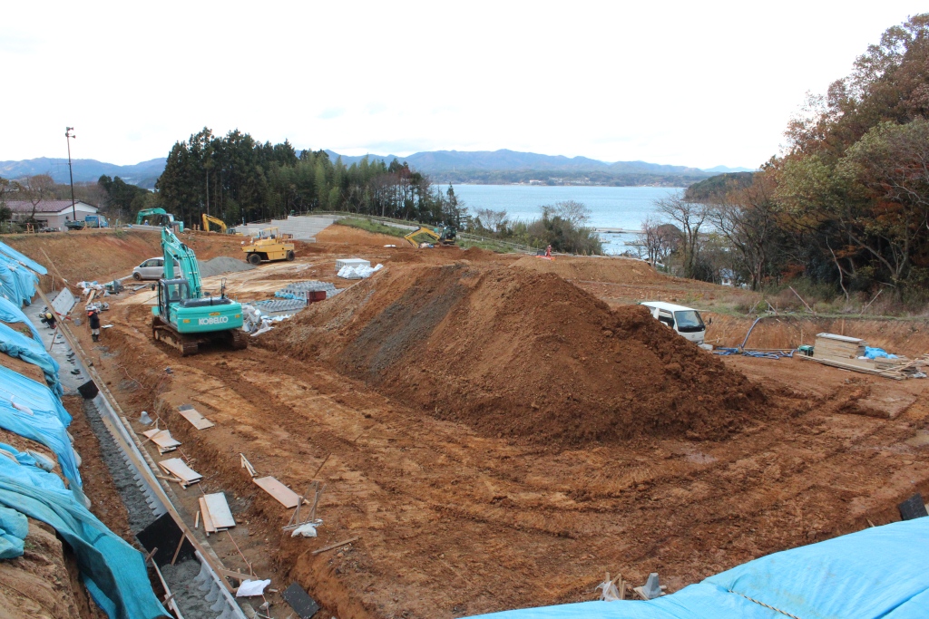 海を望む丘の造成現場で、中央に大きな土の山が盛られ、重機やトラックが配置されて作業が進められ、左側の斜面には青いシートがかけられた中、整備された道沿いに資材が並ぶ様子を写した写真