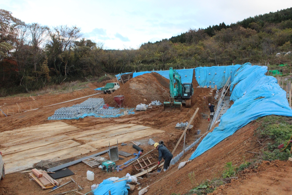 小高い斜面の造成工事現場で、右側の法面には青いシートがかけられ、中央ではショベルカーが土を掘削し、複数の作業員がブロック積みや資材の整備を行っている様子を写した写真