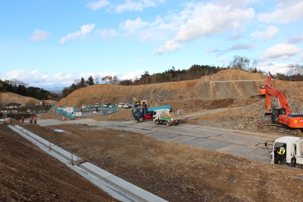 切り立った土の斜面を背景に、工事用のショベルカーやトラックが稼働しており、左奥には仮設の資材置き場や多くの車両が並ぶ整備された工事現場の写真