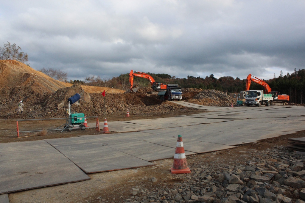 曇り空の下、広い工事現場でショベルカーがトラックに土砂を積み込む作業が行われ、舗装された仮設の通路やカラーコーン、作業員が配置されている写真