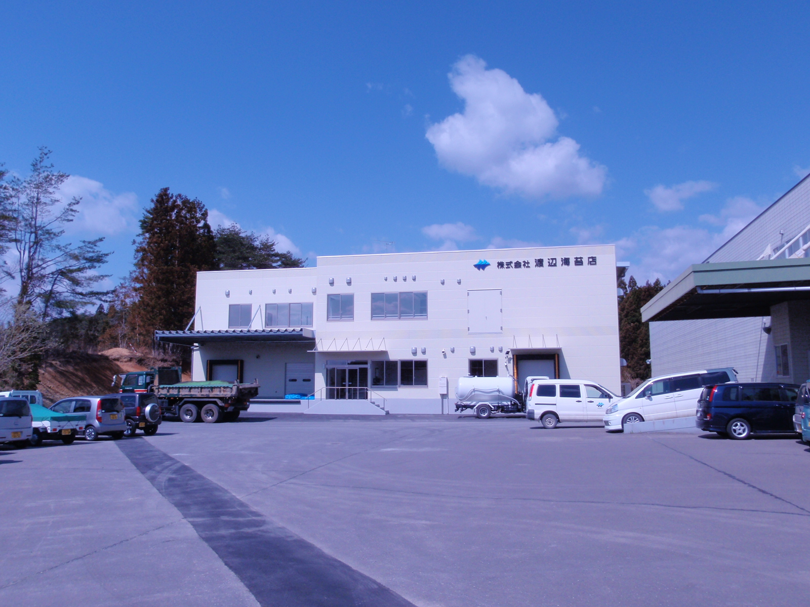 駐車スペースにトラックや数台の車が停まっている、オフホワイト色の外壁の建物の外観写真