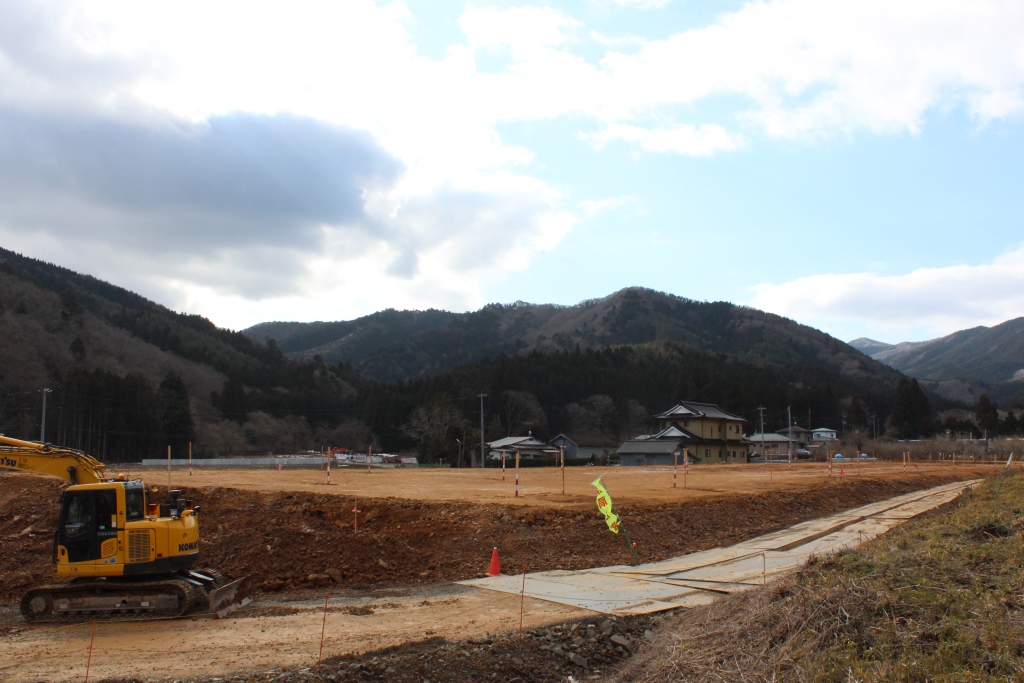 広く平らにならされた土地に工事用のショベルカーがあり、周囲には数軒の家と木々に囲まれた山々が見える写真